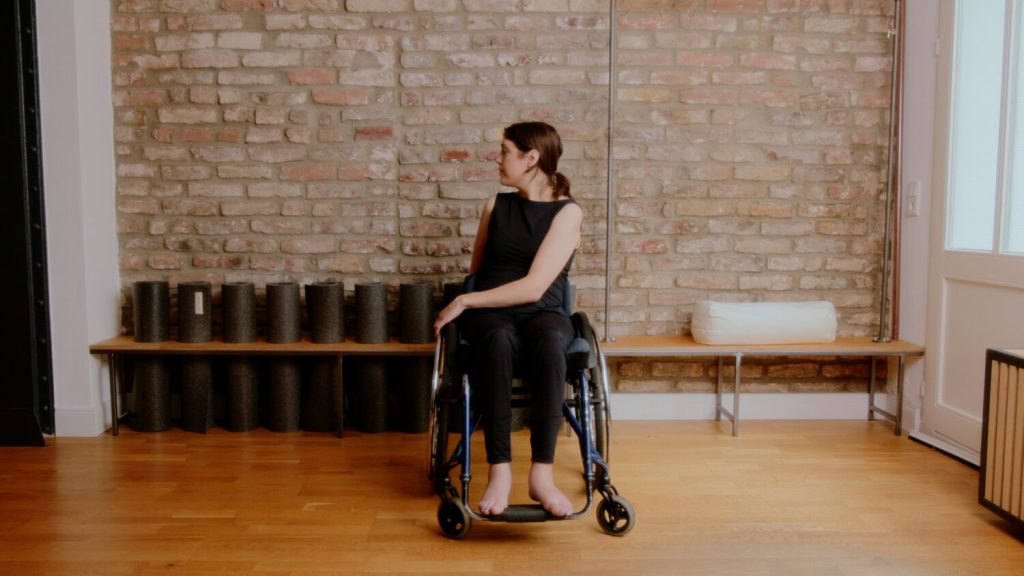 Bildausschnitt von Mechthild aus dem Yoga im Rollstuhl Video. Sie sitzt im Rollstuhl und dreht sich zu einer Seite. 